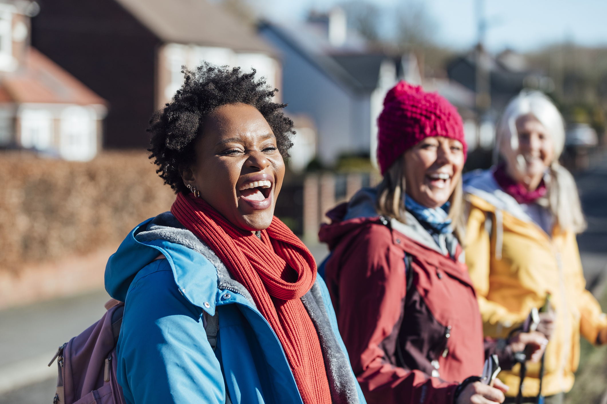 Three women laughing together on a sunlit village walk — in coats and scarves with backpacks and walking poles.
